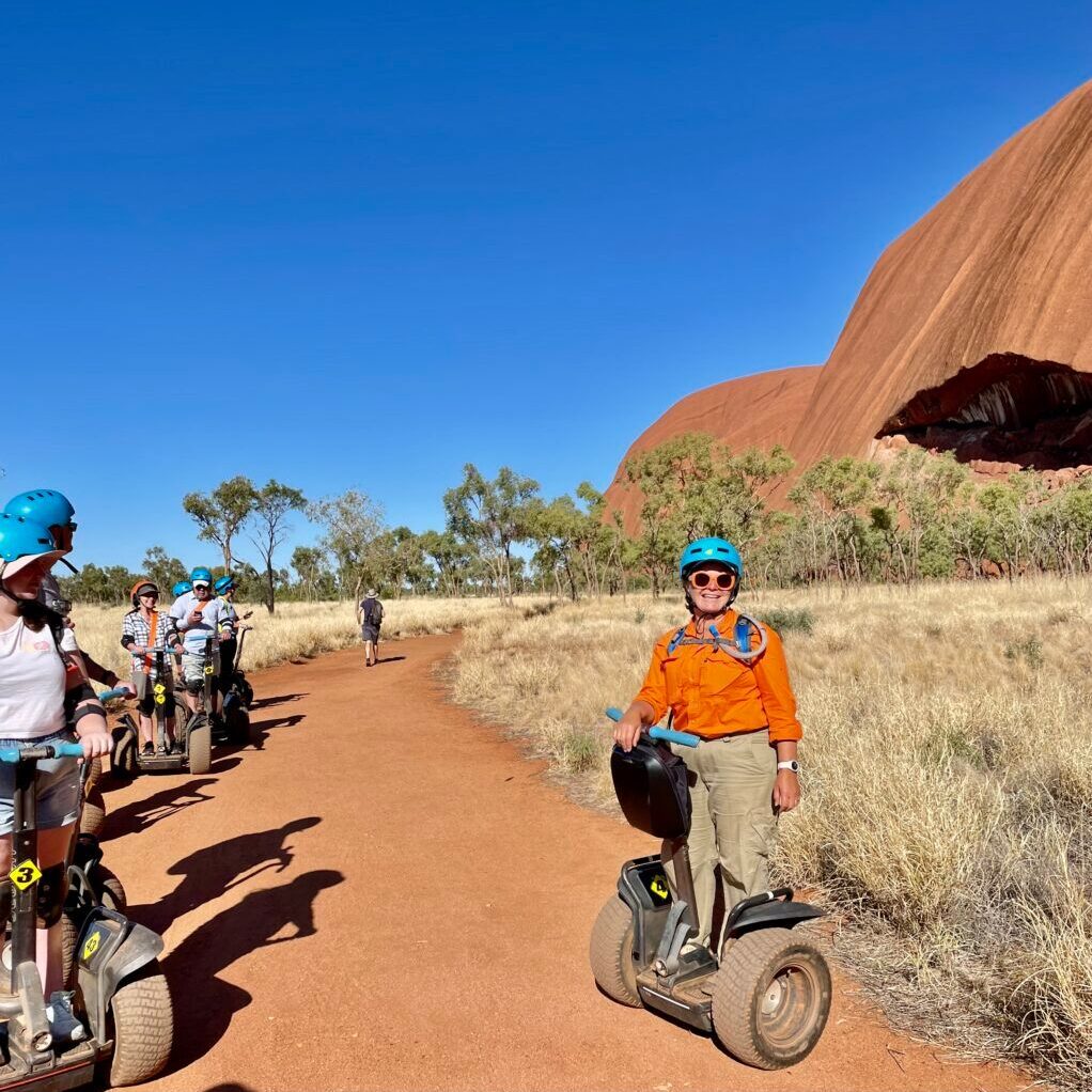 Lisa is on tour and stops with a group of Segway riders to share with them an interesting story at Uluru. Behind her Uluru is a vibrant orange and the sky is blue and appears to go on forever.